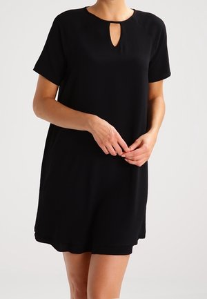 Femme portant une robe noire courte à manches courtes avec une petite ouverture en forme de goutte à l'avant, les mains délicatement croisées à la taille.