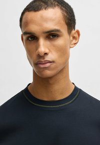 Camicetta blu navy a maniche corte con scollatura rotonda; presenta cuciture gialle in contrasto attorno al colletto e una texture morbida.