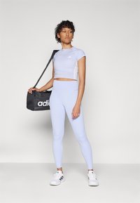 Heledad lilla lühikeste varrukatega sporditopp, millele on sobitatud kõrge vöökoha ja valgete tossudega leggingsid. Käes must adidas spordikott.