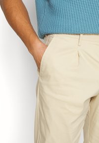 Pantaloni beige con una texture liscia e tasche laterali, abbinati a un maglione blu lavorato a maglia con motivo strutturato.