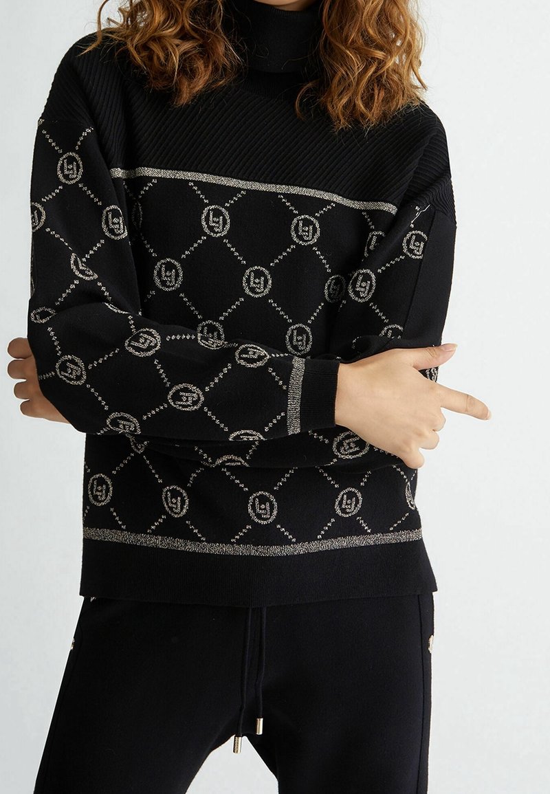Femme portant un pull noir avec des motifs géométriques et circulaires argentés, les bras croisés, une main pointant sur le côté, sur un fond uni.
