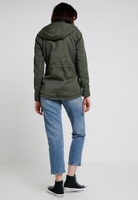 Olivengrønn, hættejakke med struktureret finish, mellemlang længde, der har en indsnævret talje. Kombineret med lyseblå, cropped jeans og sorte høje sneakers.