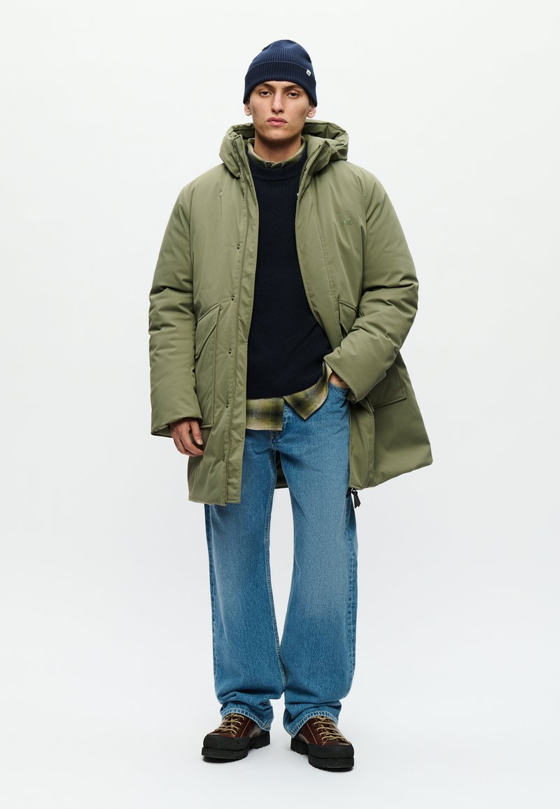 Chaqueta acolchada verde oliva con capucha, dos bolsillos frontales, llevada sobre un suéter oscuro y una camisa de franela. Jeans azules y botas marrones con cordones.