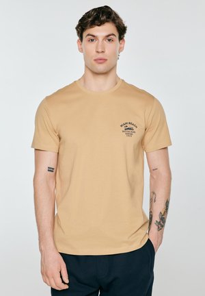 Młody mężczyzna z krótkimi kręconymi włosami, ubrany w beżowy t-shirt z grafiką Miami Beach i ciemne spodnie, stojący na jasnym tle.