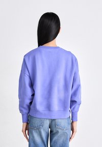 Sudadera lila con cuello redondo, mangas largas y puños de canalé; combinada con pantalones vaqueros de tiro alto en azul.