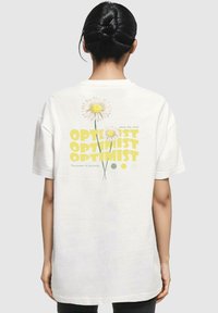 Weiße Baumwoll-T-Shirts mit einem Grafikdesign aus gelben Gänseblümchen und dem Wort "OPTIMIST" in fettgedruckter gelber Schrift auf dem Rücken. Lässiger Schnitt mit kurzen Ärmeln.