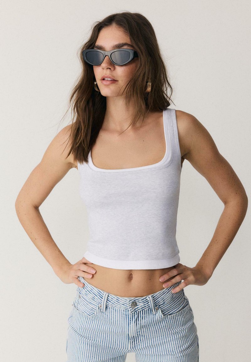 Femme aux cheveux bruns ondulés portant des lunettes de soleil noires, un haut court blanc sans manches et un jean rayé bleu et blanc, debout les mains sur les hanches.