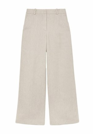 Frau trägt cremefarbene, strukturierte Hose und passenden Blazer mit einem beigen, gerippten Oberteil und goldener Anhängerkette, Hände in den Taschen.