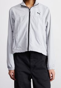 Jasnoszara kurtka zapinana na zamek z wysokim kołnierzem, elastycznymi mankietami i czarnym logo na piersi, wykonana z lekkiego materiału. Krótki krój.