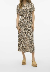 Femme portant une robe midi boutonnée en imprimé zèbre beige et noir avec une ceinture à nouer et des sandales à plateforme tressées beiges, se tenant devant un fond blanc.