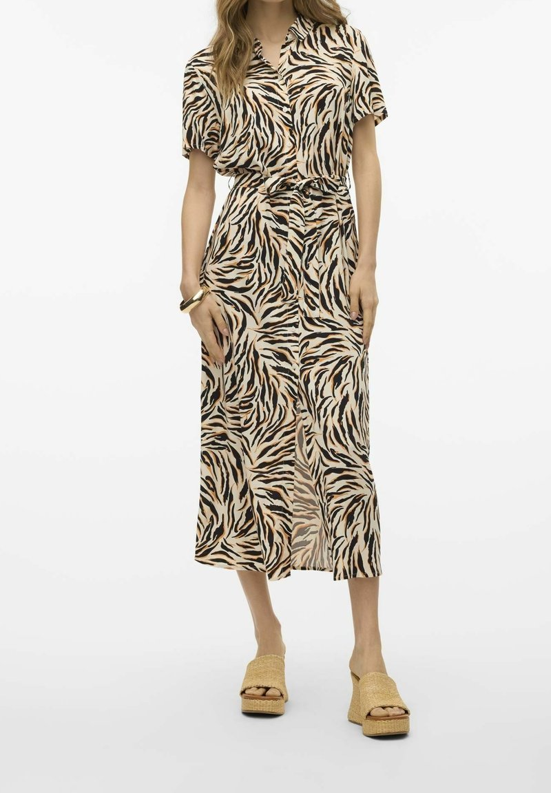 Femme portant une robe midi boutonnée en imprimé zèbre beige et noir avec une ceinture à nouer et des sandales à plateforme tressées beiges, se tenant devant un fond blanc.