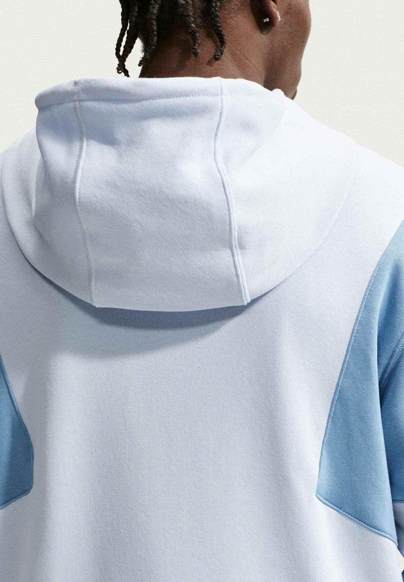 Persoon met korte twists draagt een witte hoodie met lichtblauwe schouderpanelen, van achteren getoond tegen een effen, lichte achtergrond.