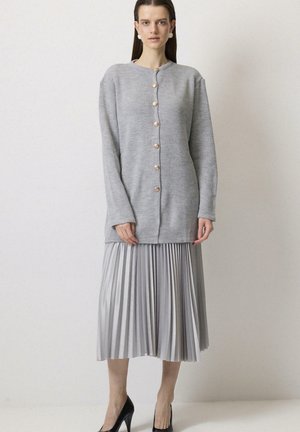 Vrouw draagt een lichtgrijze gebuttonde cardigan over een zilveren geplooide midi-rok met zwarte hoge hakken, staand tegen een effen witte achtergrond.