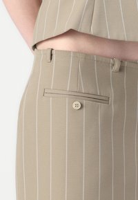 Beige stripete bukser med en glatt tekstur, som har en baklomme med en rund knappelukking og beltehemper.