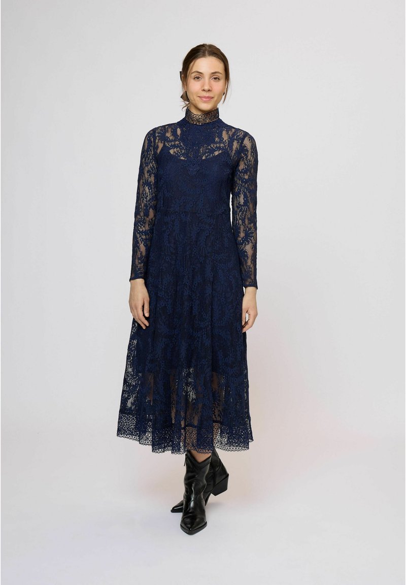 Tmavomodré čipkované šaty s dlhými rukávmi, vysokým golierom a vrstvenou sukňou. Nosené s čiernymi členkovými topánkami, ktoré majú textúrovaný vzor a priehľadný materiál.