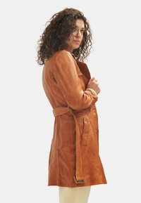Tan leather trench coat with a belted waist, side pockets, button closures, and a slightly textured surface; mid-thigh length.