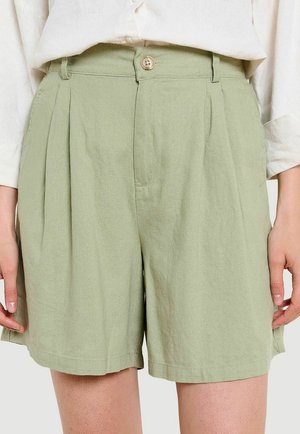 Pantalones cortos de talle alto en verde claro, entallados, con pliegues frontales, cierre de botón y cremallera, combinados con una camisa de manga larga color crema.