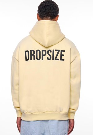 Lys gul hettegenser med stor svart "DROPSIZE" trykk på baksiden. Har hette, løs passform og ribbestrikkede mansjetter og hem.