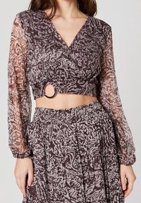 Blusa trasparente a maniche lunghe con scollatura a V profonda e vita sottile, con accento circolare. Il tessuto è a fantasia in tonalità sobrie di marrone.