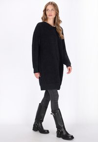 Vrouw wandelt in een zwarte lange fuzzy trui, grijze skinny jeans en zwarte leren kniehoge laarzen met gespen.