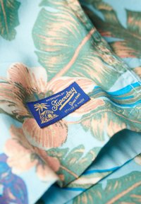 Camicia in stile hawaiano con uno sfondo azzurro chiaro, decorata con motivi floreali rosa e verdi. Include un'etichetta blu con la scritta "Superdry".