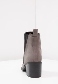 Grijze suède enkelboot met elastische zijpanelen, een korte blokhak en een lus aan de achterkant. Getextureerde zwarte accenten langs de zijkanten.
