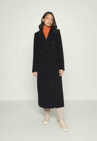 Woman wearing a long black double-breasted coat, orange turtleneck sweater, beige pants, and beige ankle boots, standing on a plain background.