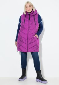 Žena v fialové vzorované bezrukávové kapuci vestě přes modrý svetr, tmavých džínách a černých botách s fialovými podešvemi, stojící na bílém pozadí.