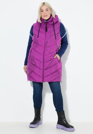 Vrouw in een paarse gestippelde mouwloze hoodie-vest over een blauwe trui, donkere jeans en zwarte laarzen met paarse zolen, staande tegen een witte achtergrond.