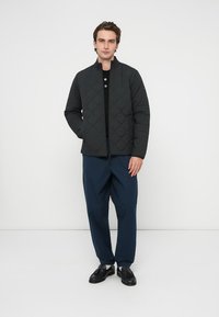 Quilted black jacket with a stand collar, zip closure, and navy pants. Model stands on a plain background, wearing black shoes.