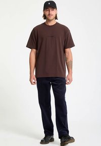 Brown cotton t-shirt with crew neckline and front text detail, paired with dark navy corduroy pants and black shoes. Matching cap on head.