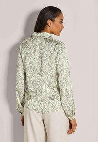Vrouw met donker haar draagt een lichtgroene blouse met paisleypatroon en lichtbeige broek, staand tegen een effen muur.
