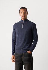 Navy zippered pullover sweater with high collar, knit texture, and ribbed cuffs. Paired with dark jeans featuring visible stitching details.