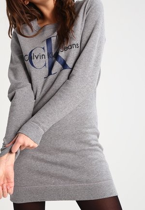 Osoba ubrana w szarą sukienkę z długim rękawem z niebieskim logo "Calvin Klein Jeans", poprawiająca rękaw, stojąc na jednolitym tle.