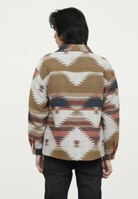 Wool button-up shirt with a geometric pattern in earthy tones of brown, cream, and red, featuring a collar and cuffed sleeves.