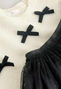 Cremfarbener Strickpullover mit drei schwarzen Stoffschleifen, kombiniert mit einem schwarzen Netzrock mit gepunktetem Muster und elastischem Bund.