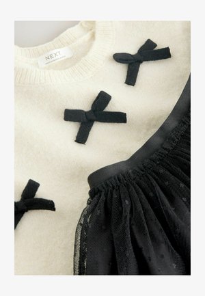 Krämfärgad stickad tröja med tre svarta tygrosetter, kombinerad med en svart meshkjol med prickigt mönster och elastisk midja.