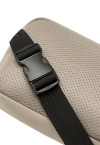 Beige vadderad yta med nätstruktur och svart nylonrem med en plastspänne. Rund form med mjuk kant.