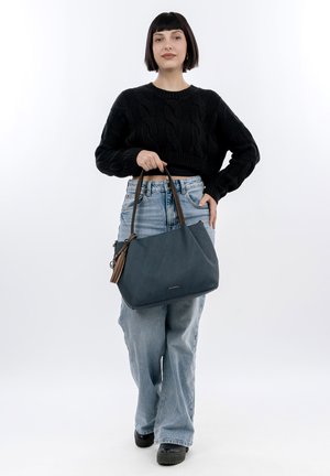 Donna con maglione nero e jeans che tiene una grande borsa blu scuro con manici marroni, in piedi davanti a uno sfondo bianco.