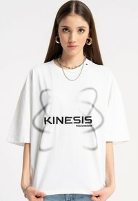 Młoda kobieta w białej, oversizedowej koszulce z napisem "KINESIS subconscious" oraz abstrakcyjnymi szarymi kształtami, uzupełniona srebrnymi łańcuszkami i kolczykami w kształcie kół.
