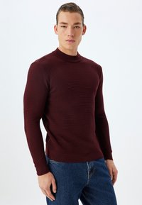 Bordeaux, gestructureerde trui met een hoge hals en lange mouwen, gecombineerd met blauwe jeans. Het ontwerp heeft een ribbelpatroon over het geheel.