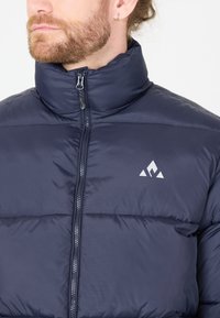 Veste rembourrée bleu marine avec un col haut, fermeture éclair à l'avant et un petit logo blanc sur la poitrine. Texture du tissu lisse et léger.