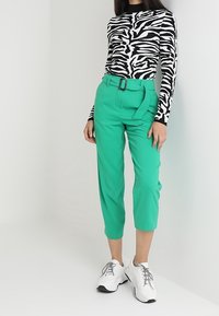 Osoba ubrana w bluzkę z długim rękawem w wzór zebry, zielone spodnie o wysokim stanie i ortezie, oraz białe chunky sneakersy, stojąca na jednolitym tle.