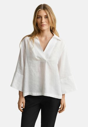 Femme aux longs cheveux blonds portant un chemisier blanc ample à col en V avec des manches larges et un pantalon noir, debout devant un fond uni.