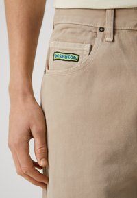 Bež jeans hlače z vidno šivanko, sprednjim žepom z večbarvno vezeno oznako DC Shoe Co. in gladko teksturo.