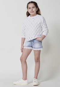 Un pull en maille blanche avec un motif en losanges texturé, associé à un short en denim avec des bordures en dentelle blanche. Des baskets blanches complètent la tenue.
