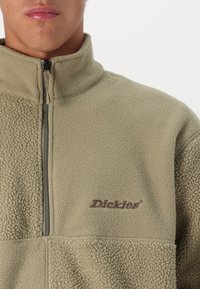 Olivinės žalios vilnonės švarkas, su pusiau užtrauktuku, tekstūruota paviršiumi ir ruda siuvinėta logo kairėje krūtinės pusėje.