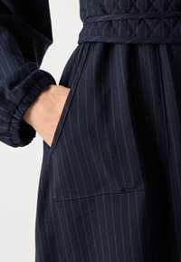 Main dans la poche d'un vêtement bleu marine à fines rayures avec une ceinture matelassée et un poignet élastique sur la manche.