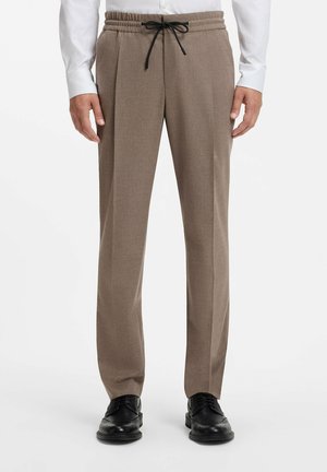 Mand iført beige bukser med lige ben med elastisk talje og sort snor, kombineret med sorte pæne sko og en hvid skjorte.