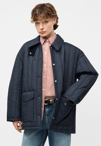 Νεαρό άτομο που φοράει μπουφάν ναυτικού μπλε με επένδυση πάνω από κόκκινο πουκάμισο με ρίγες και μπλε τζιν, με το ένα χέρι στην τσέπη του μπουφάν.